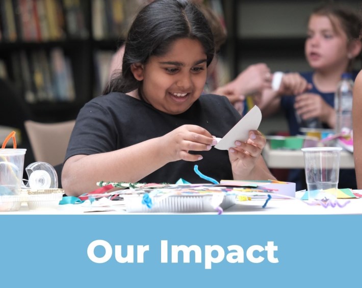 Young girl sitting at a table working on a craft project. She is smiling. The words read Our Impact underneath. This image links to our Impact Report