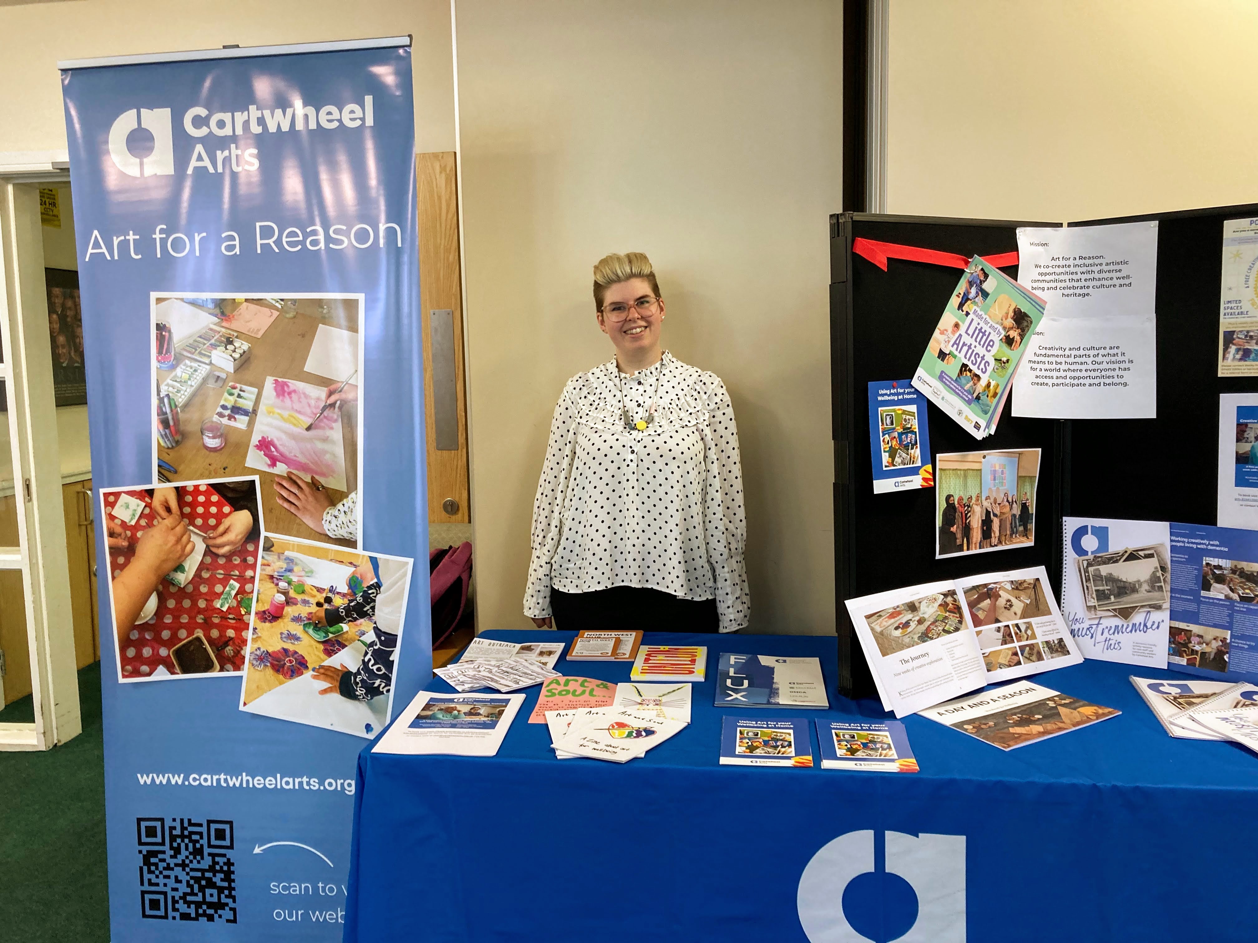 Becky standing behind a Cartwheel Arts events table with flyers, leaflets and banners. She is smiling 