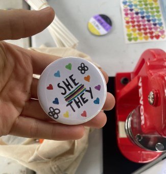 This image shows a hand holding a badge which reads she/they and is decorated with different coloured hearts