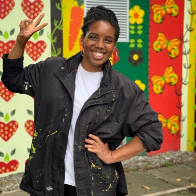 This image shows a lady smiling with her hand on her hip and doing the peace sign behind her is a decorated wall of hearts, flowers and bows